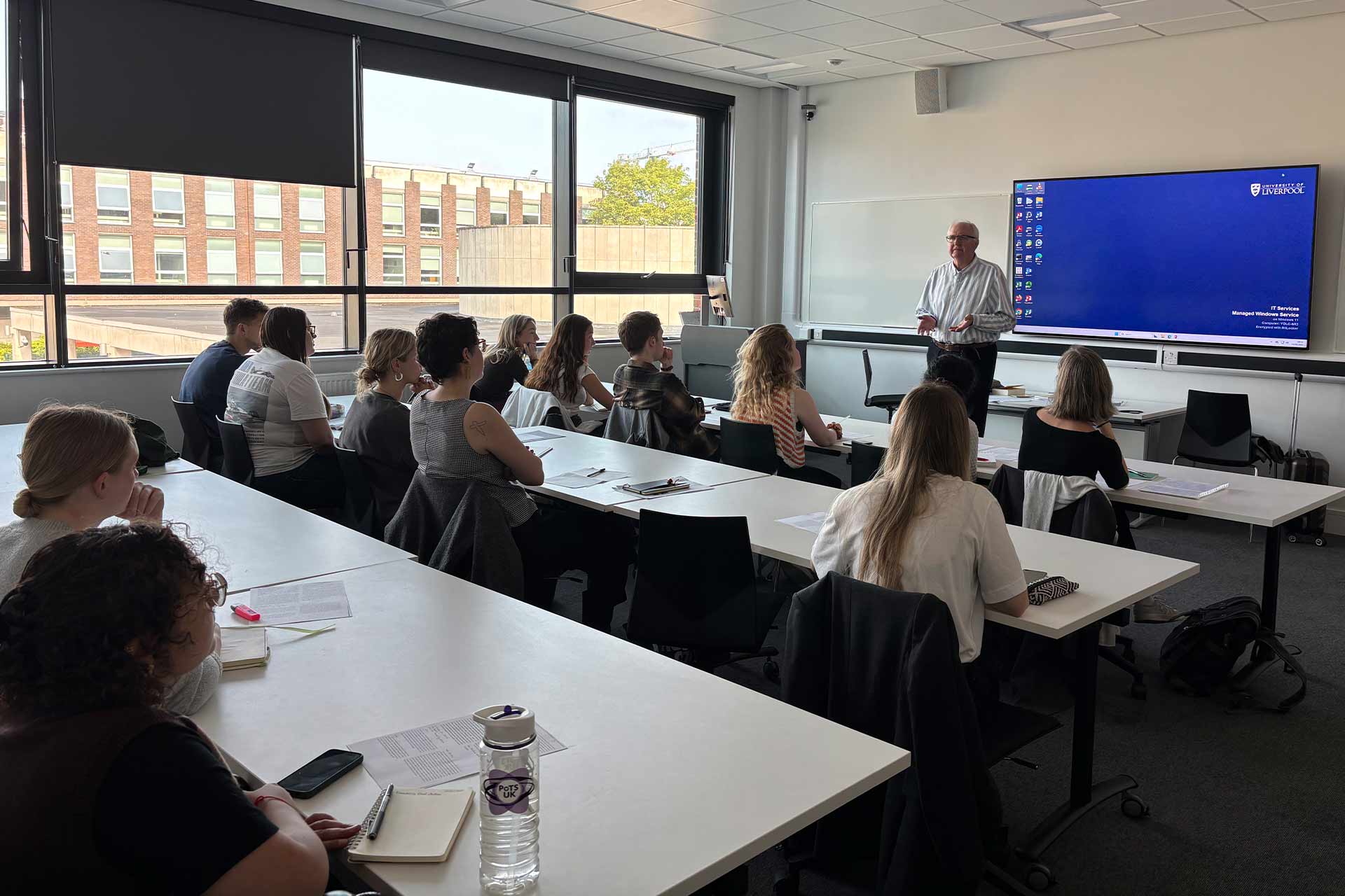 A teaching room with students and a person at the front presenting with a screen behind them.