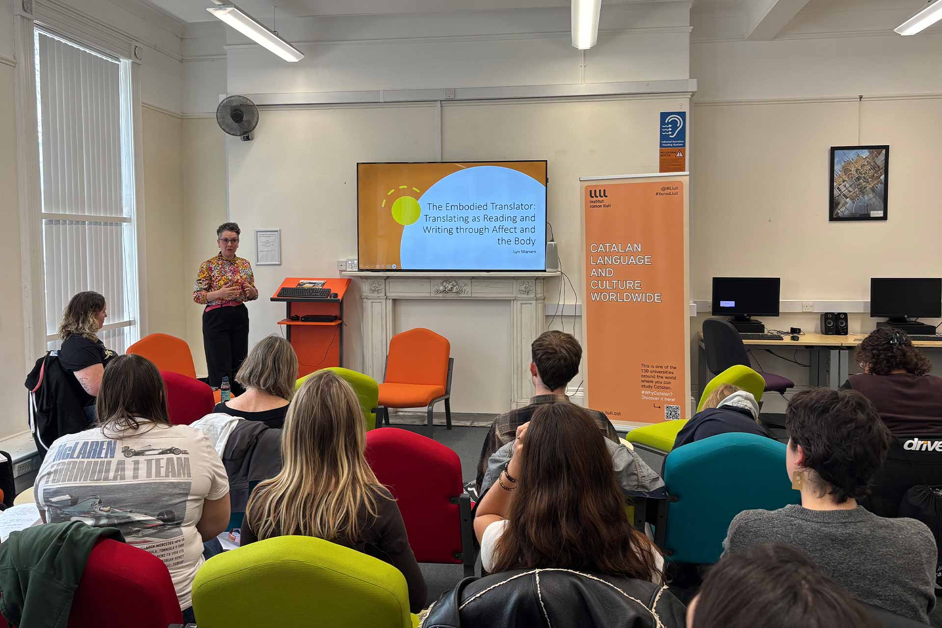 A group of people sitting and listening to a person presenting at the front, whilst they stand in front of a powerpoint slide that reads 'The embodied translator: translating as reading and writing through affect and the body'