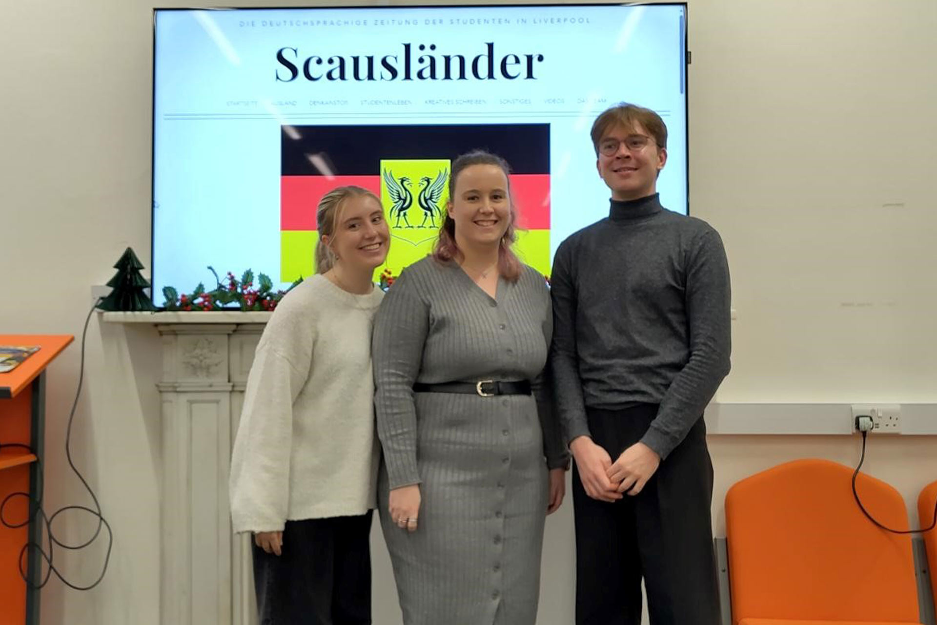 Three students smiling and standing in front of a presentation on a projector screen that says 'Scauslander' with a German flag below it.