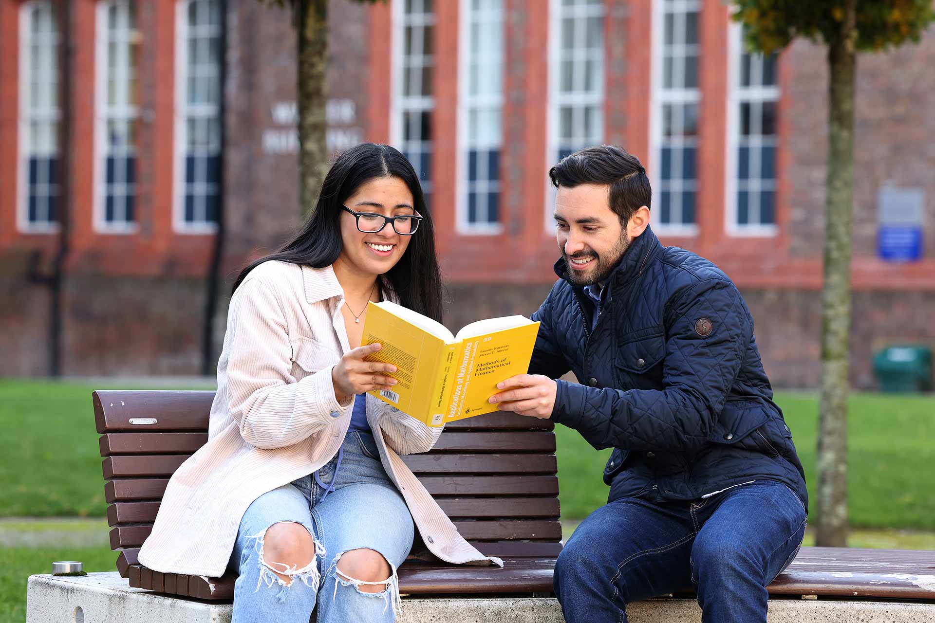 Two students study a textbook while sat on a bench in the Quad.