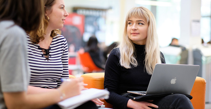 A photograph of two postgraduate students chatting. One is holding a pen and paper, and the other has a laptop.