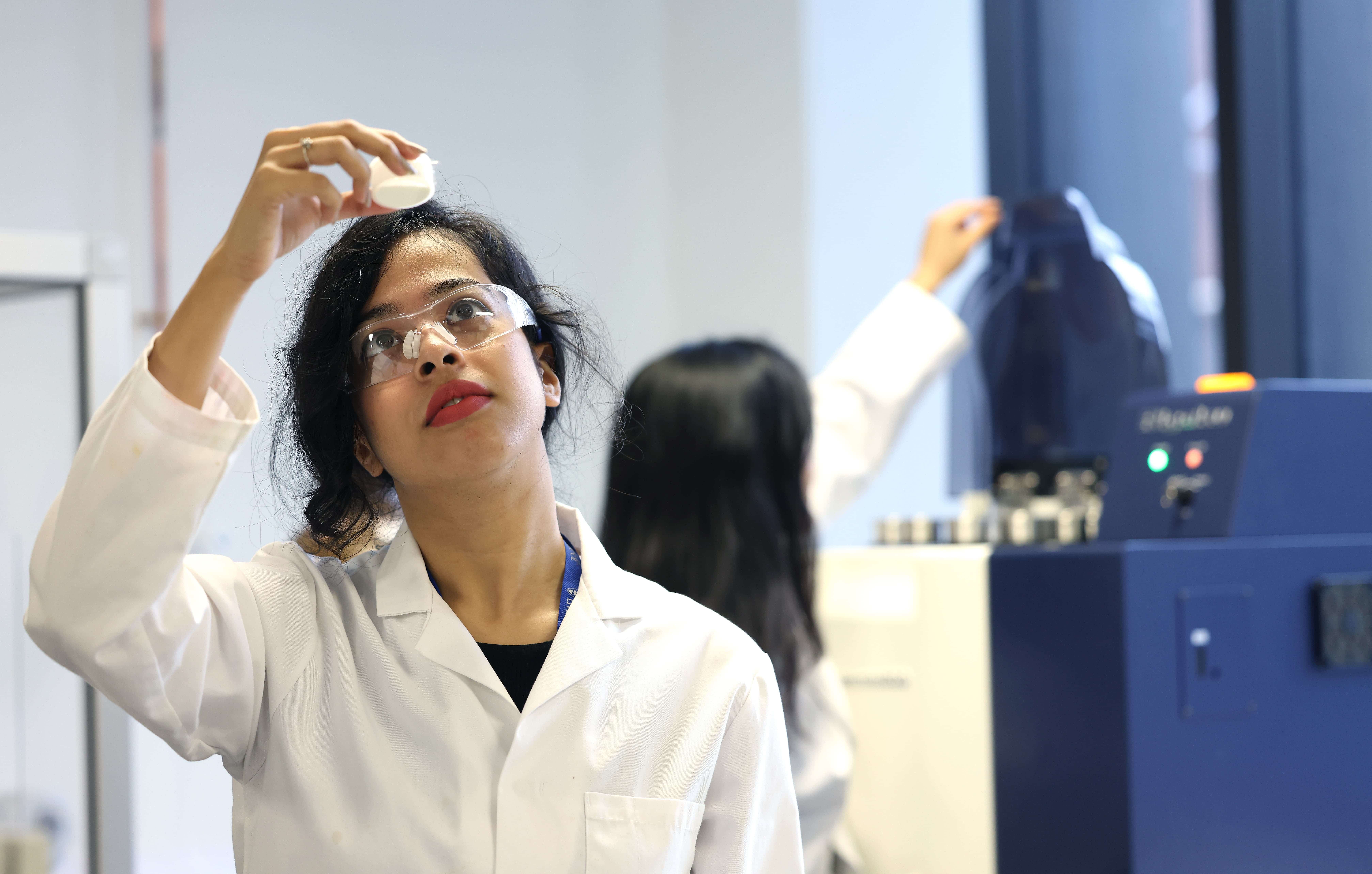 A postgraduate research student holding up a piece of equipment in a lab, wearing a lab coat and safety goggles.