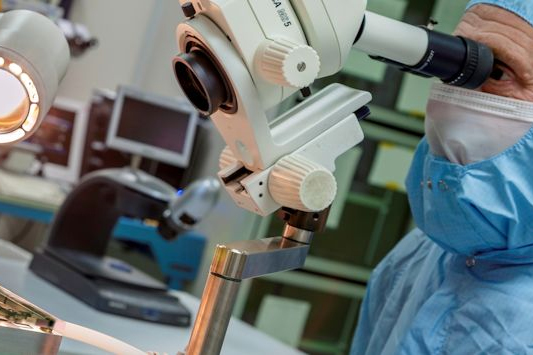 A researcher in blue scrubs and a face mask inspects a sample through a microscope.