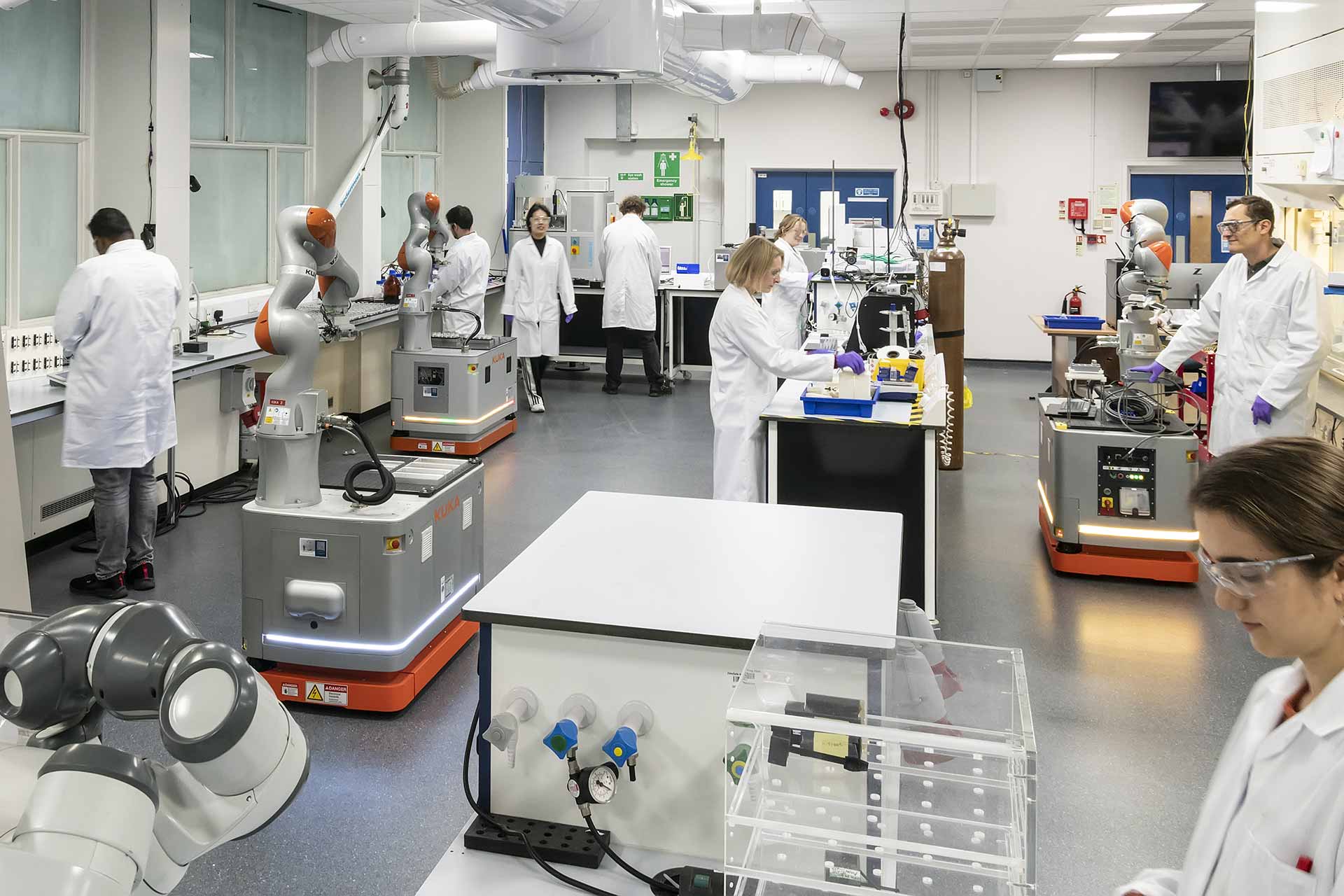 Scientists in a modern lab work alongside robotic arms and automated equipment at various workstations.