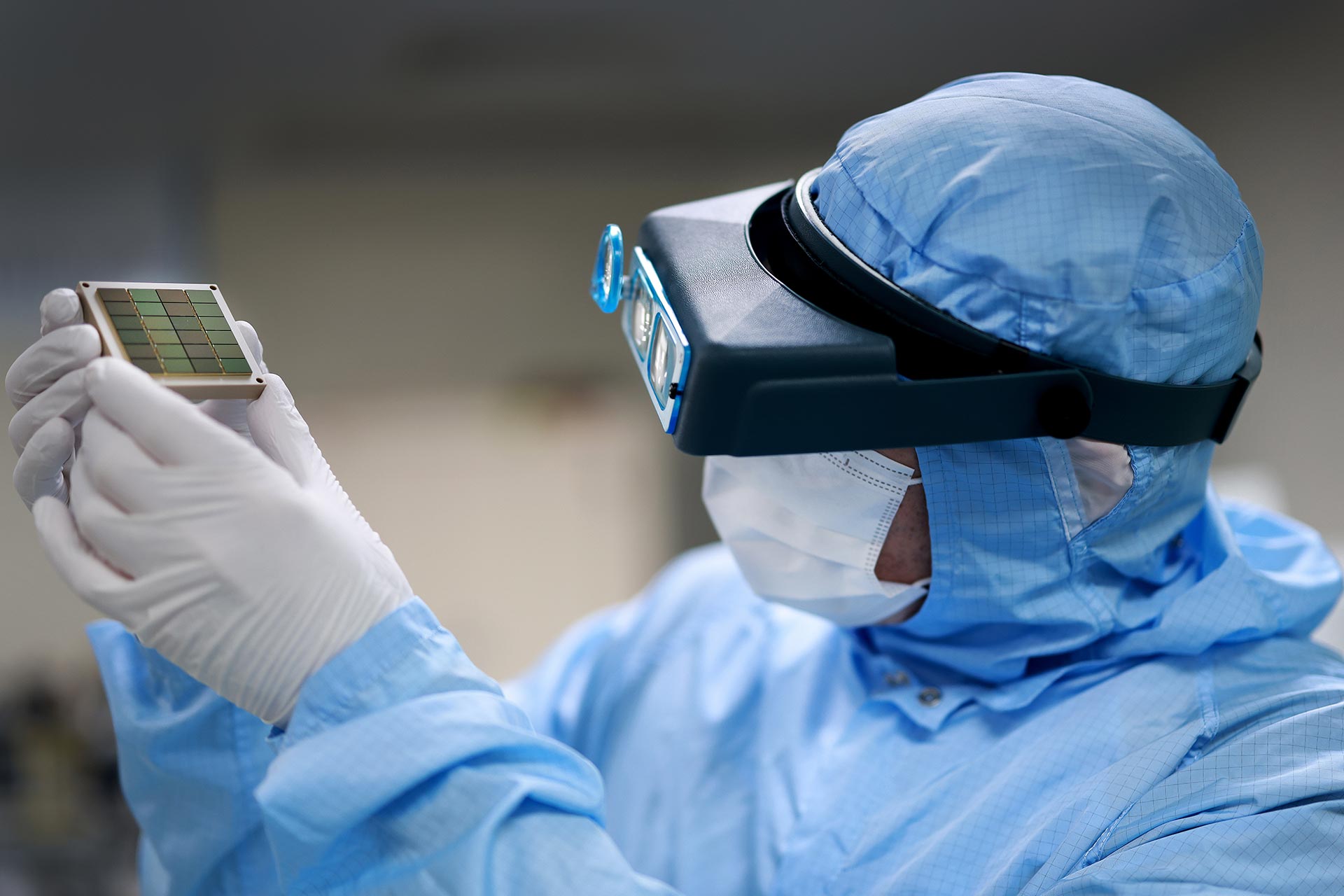 A researcher dressed in blue scrubs and wearing a futuristic eye mask inspects a lab sample.
