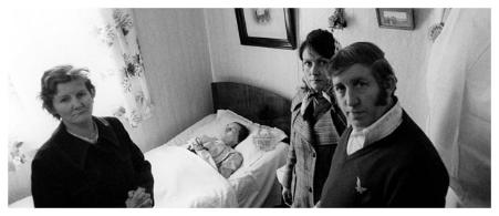 Three people standing around a dead body lying in a bed