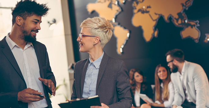 Two people standing talking with a large world map on the wall in the background