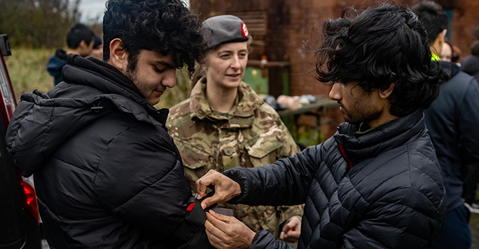 Two male student doctors stood either side of a female Army member. One student is dressing the others arm with a battle tourniquet, whilst the Army Officer observes.