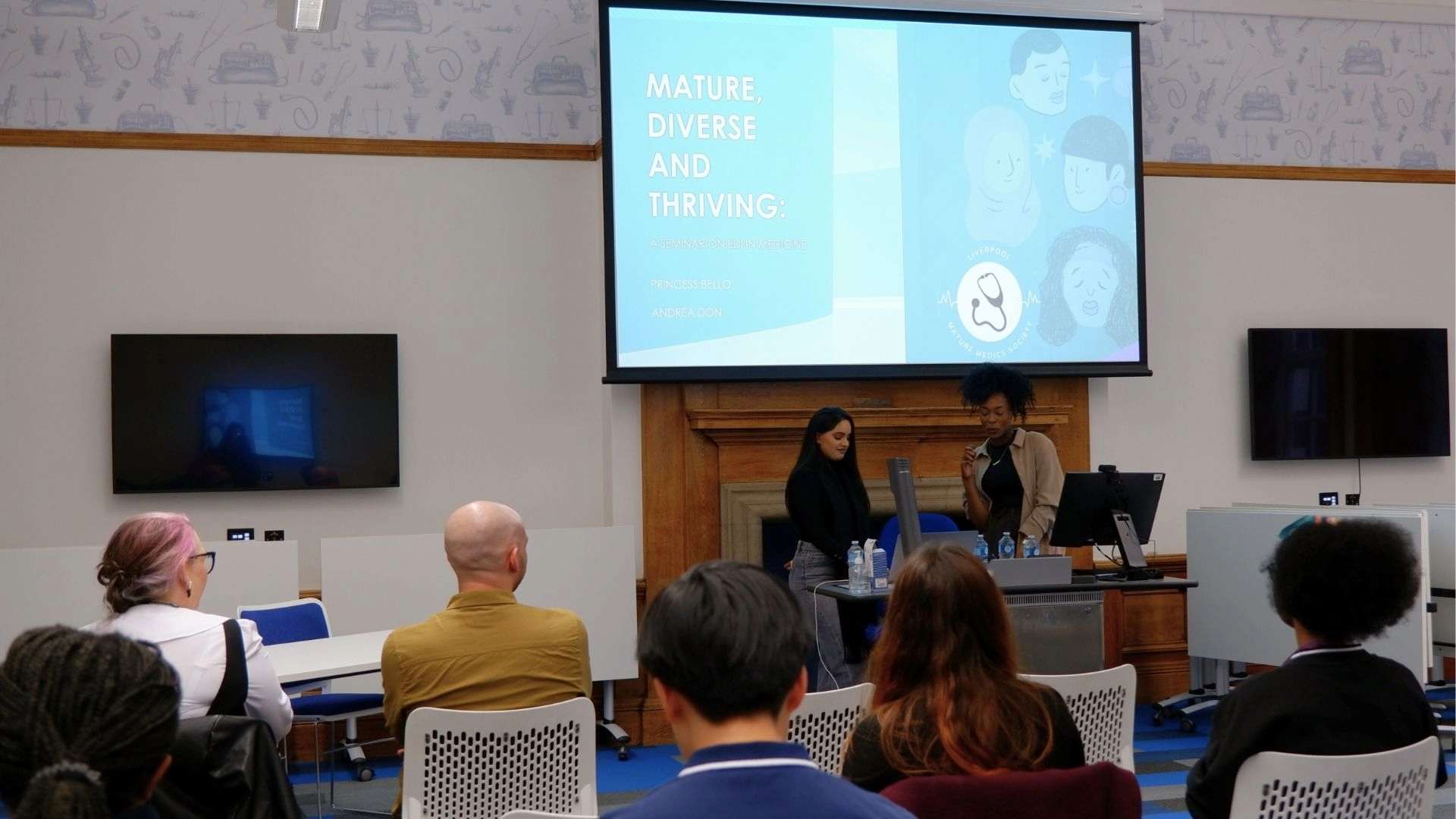 Two presenters stand at the front of a lecture room, speaking to an audience seated in rows. A large screen behind them displays the title “Mature, Diverse and Thriving: A Seminar on Identity in Medicine” as part of a Liverpool Mature Medics Society event.