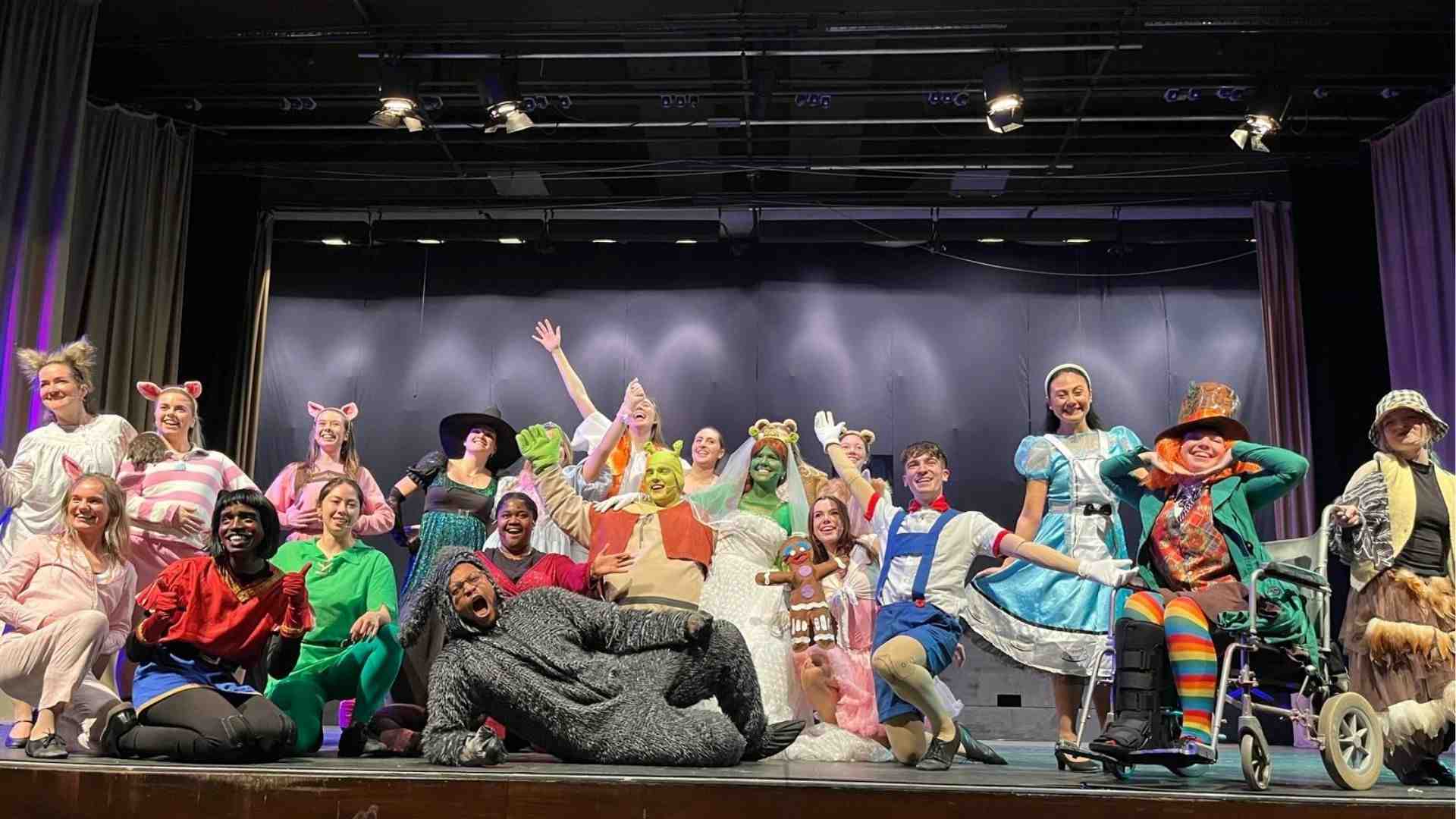 Group photo of the full cast in costume, smiling and posing together on stage.