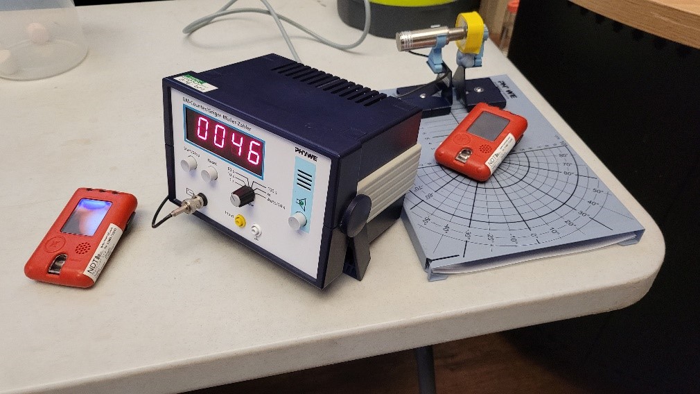 Our Indium Source (Yellow case) next to the GM tube (Silver cylinder) connected to the counter (Blue box with numbers). We also had ‘Dosimeters’ (Orange devices) to measure how much or a radiation dose someone may get at different distances