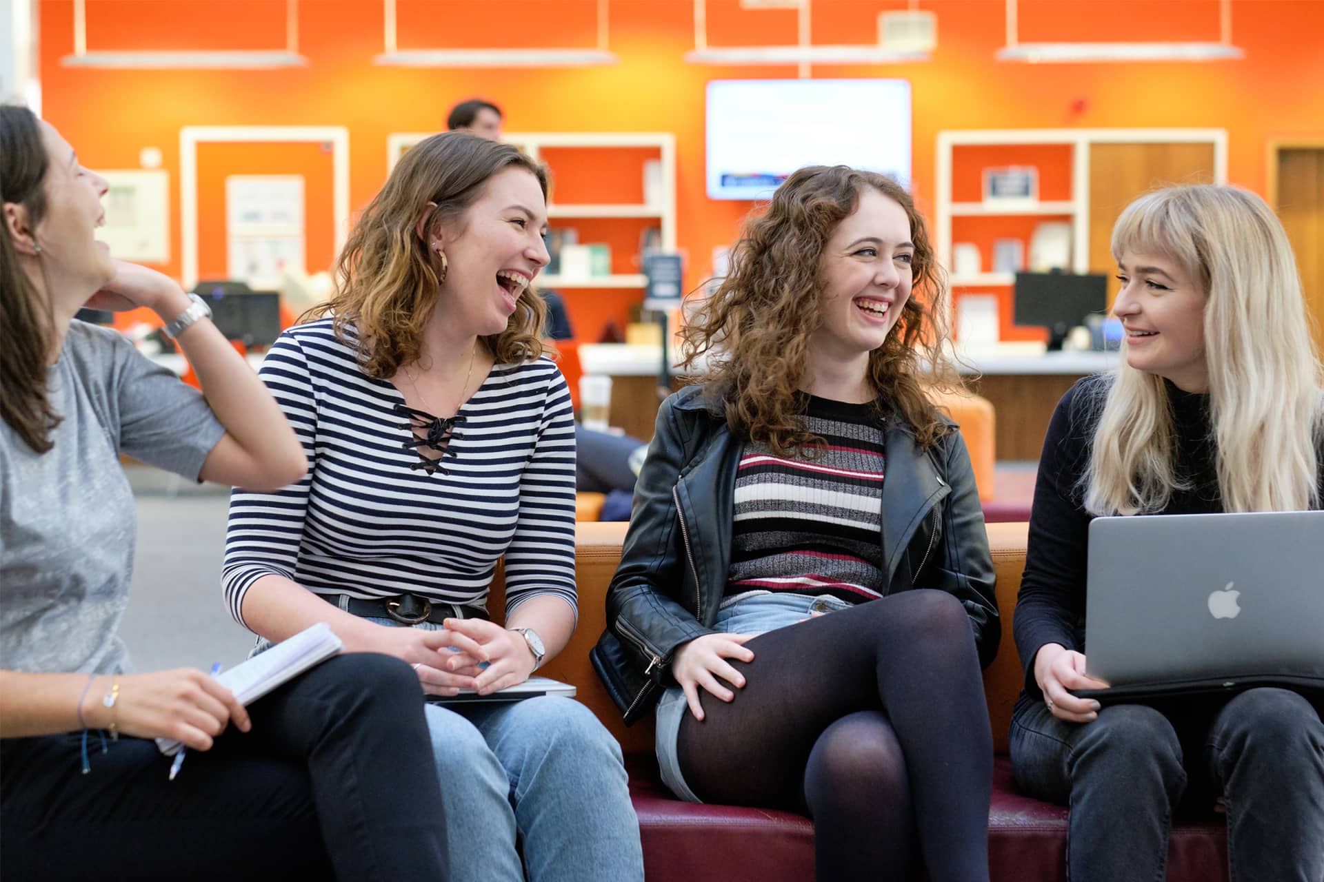 Four students sit on a couch smiling and ooking at eachother.