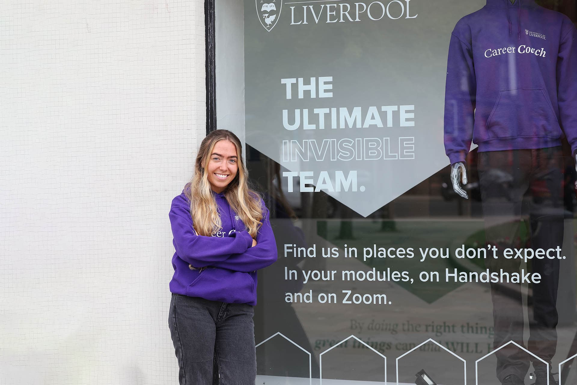 A career coach stands next to the Universitys career centre. She is smiling and looks ready to help.